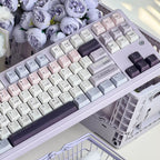 A pastel mechanical keyboard with a lavender chassis and matching keycaps in soft pink, cream, light blue, and dark purple, styled on a desk with faux flowers and a plastic crate.