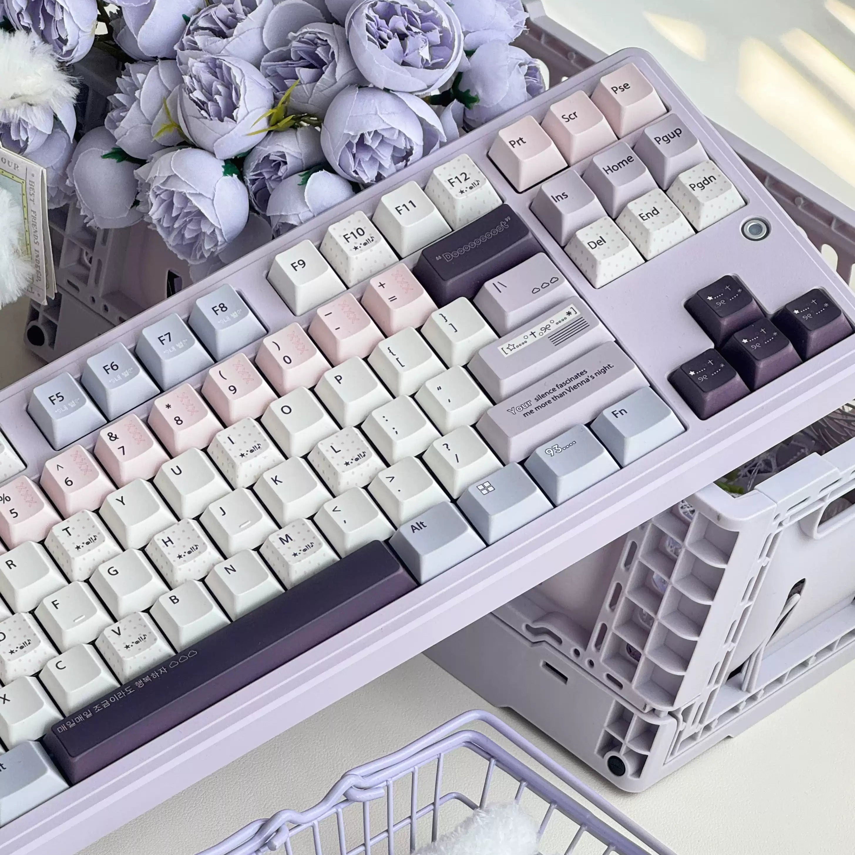 A pastel mechanical keyboard with a lavender chassis and matching keycaps in soft pink, cream, light blue, and dark purple, styled on a desk with faux flowers and a plastic crate.