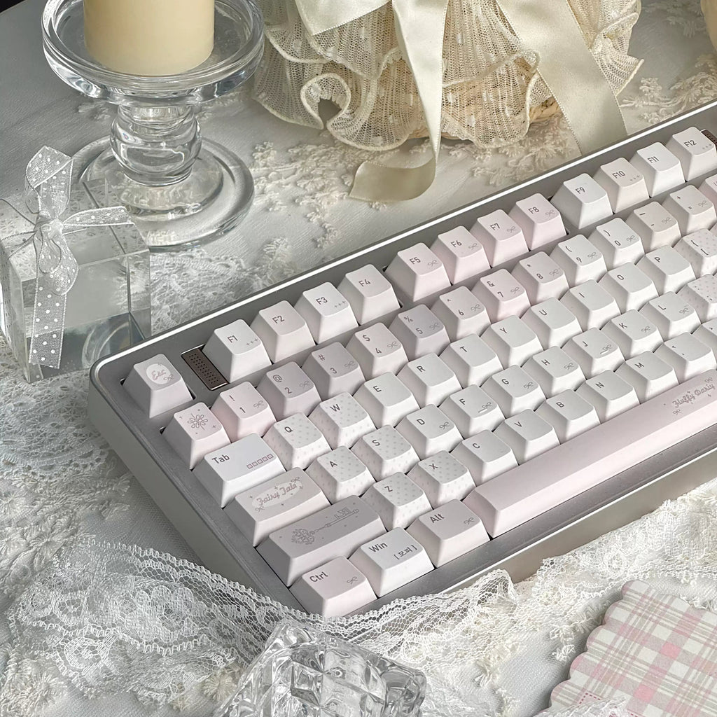 A mechanical keyboard with pale pink and white keycaps and a silver-gray frame, styled on a white lace surface with a candle, gift box, and ribbon in the background.