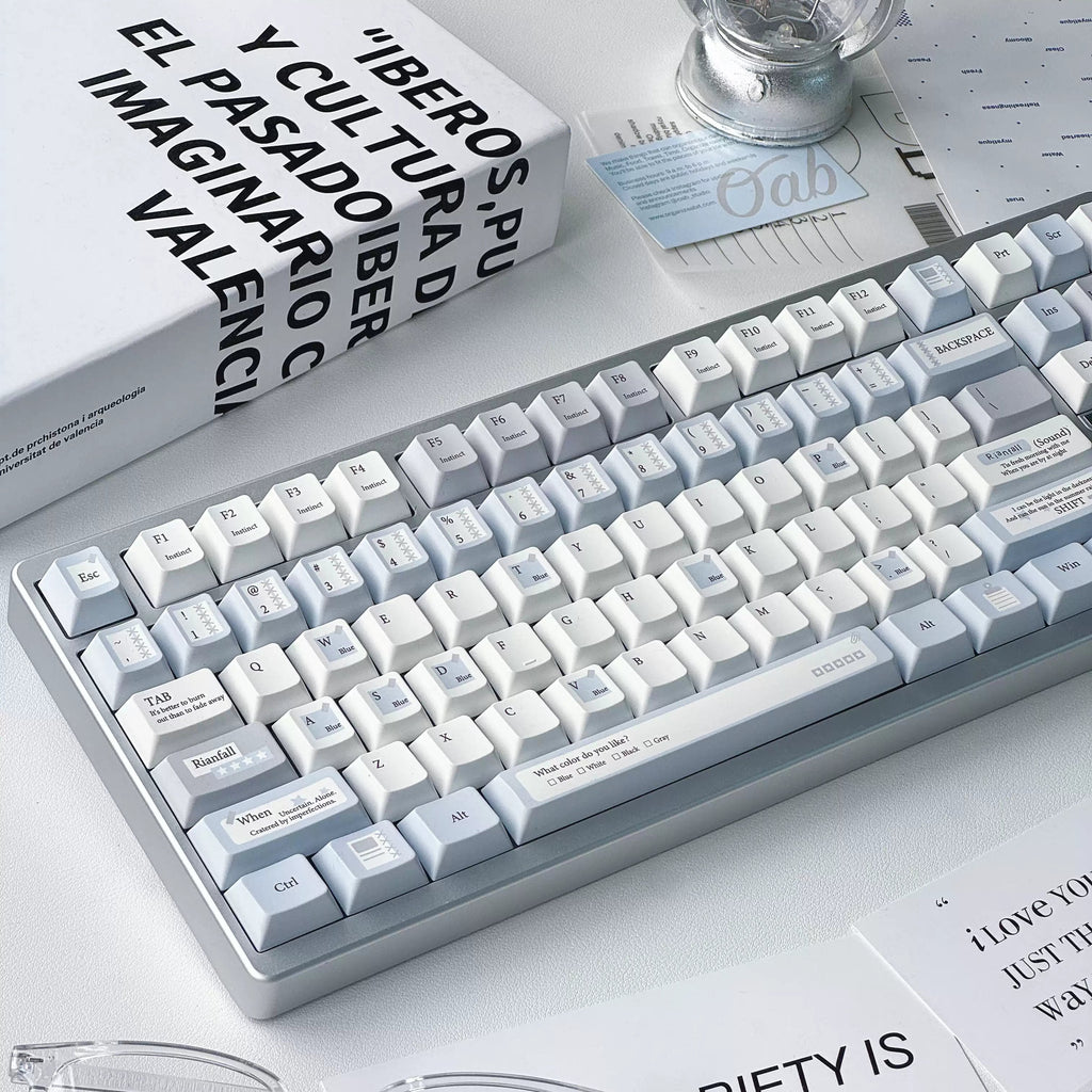 A clean, modern mechanical keyboard with white and light gray keycaps and a silver-gray frame, styled on a desk with glasses, a book, and a light bulb.