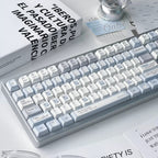 A clean, modern mechanical keyboard with white and light gray keycaps and a silver-gray frame, styled on a desk with glasses, a book, and a light bulb.