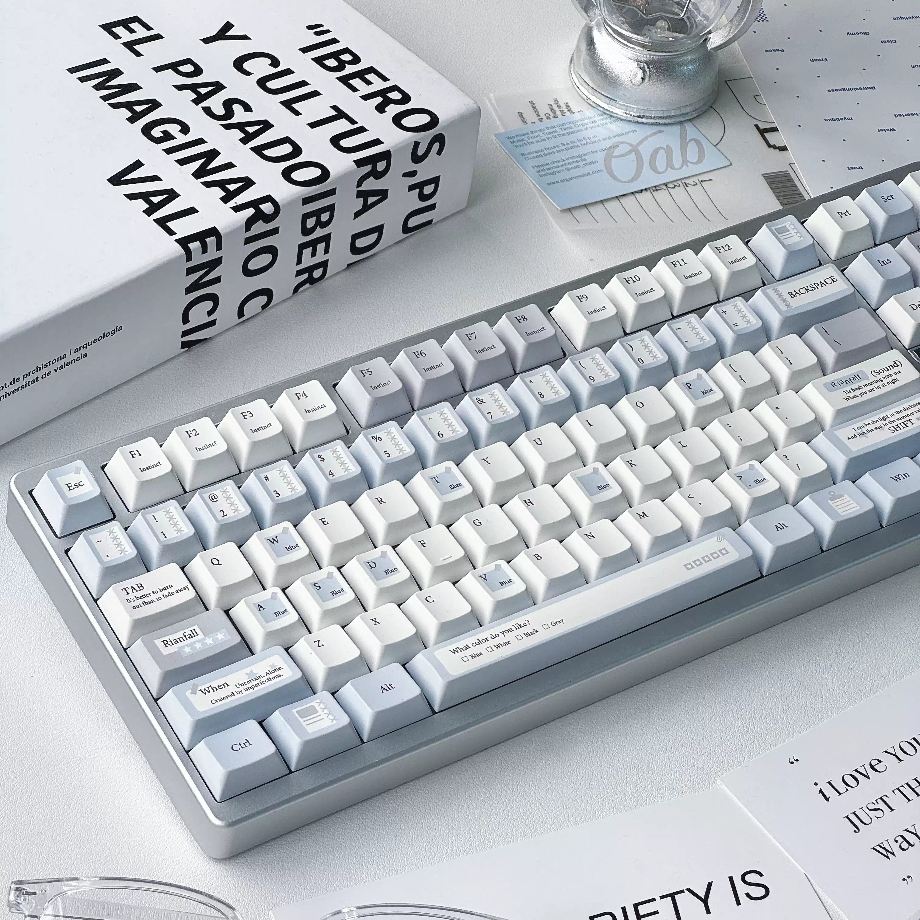 A clean, modern mechanical keyboard with white and light gray keycaps and a silver-gray frame, styled on a desk with glasses, a book, and a light bulb.