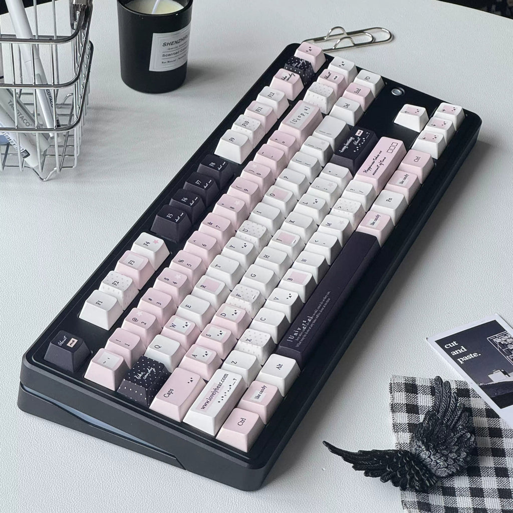 A mechanical keyboard with a black frame and pastel pink, white, and dark gray keycaps, styled on a white desk with a candle, wire basket, and small accessories.