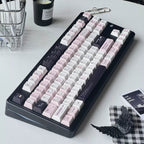 A mechanical keyboard with a black frame and pastel pink, white, and dark gray keycaps, styled on a white desk with a candle, wire basket, and small accessories.