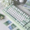 A pastel green and white mechanical keyboard styled on a tabletop with white flowers, a white lamp, and small landscape cards.
