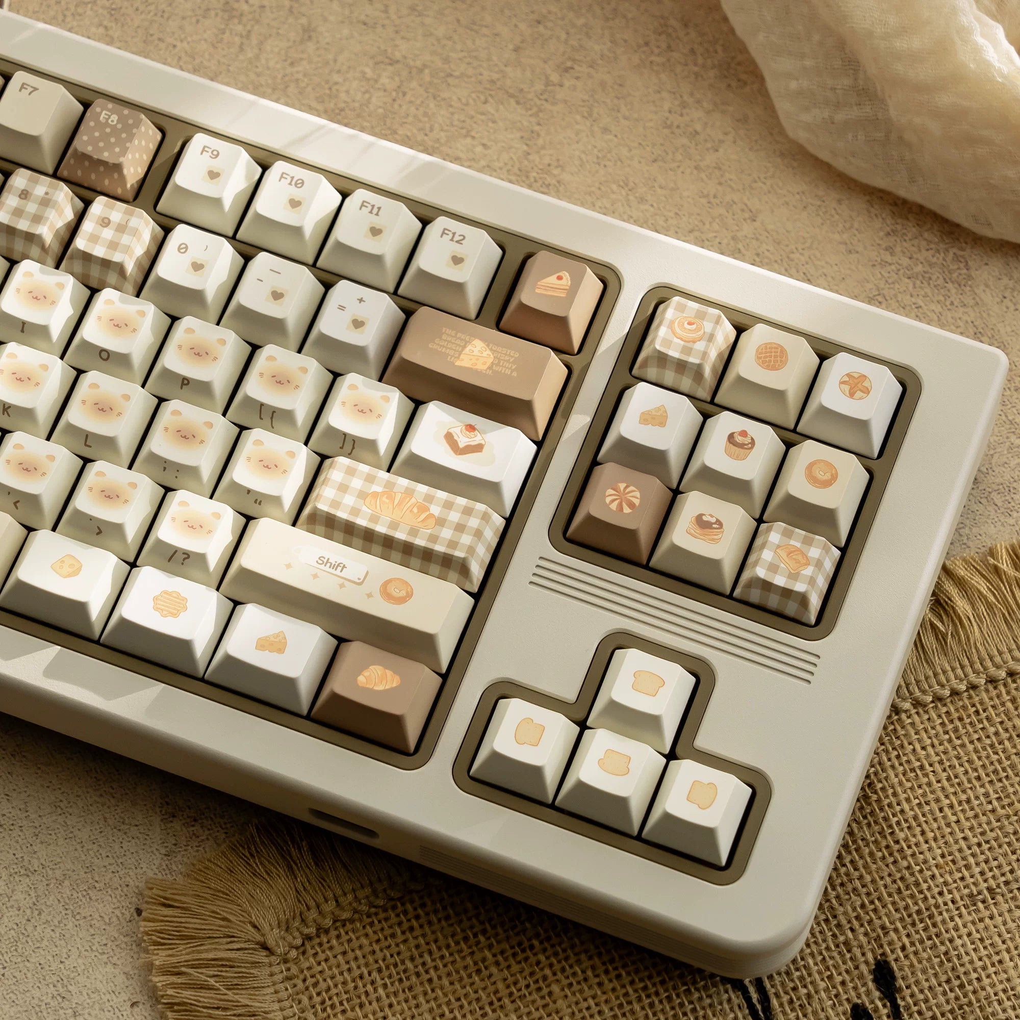 A close-up of a mechanical keyboard with cream, beige, and light brown keycaps decorated with cute food illustrations, set on a textured beige fabric background.
