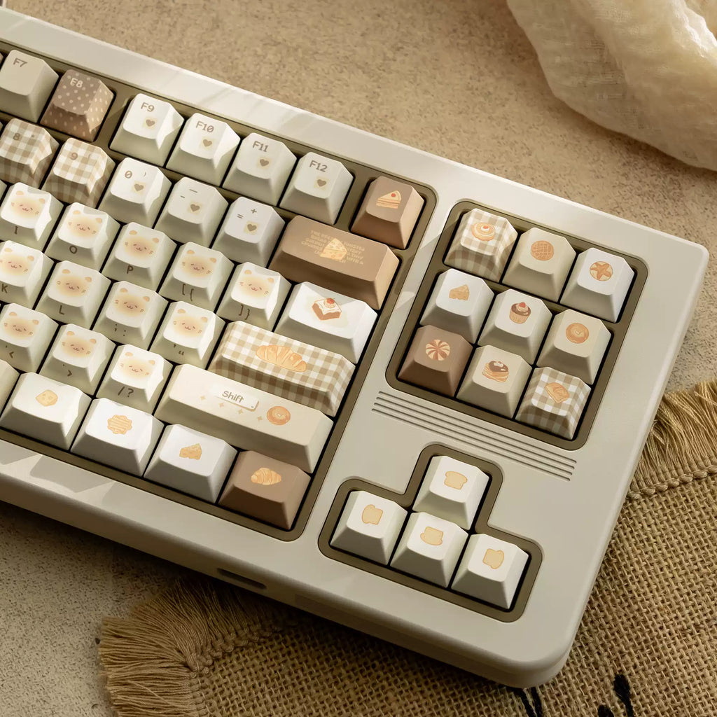 A close-up of a mechanical keyboard with cream, beige, and light brown keycaps decorated with cute food illustrations, resting on a textured beige fabric surface.