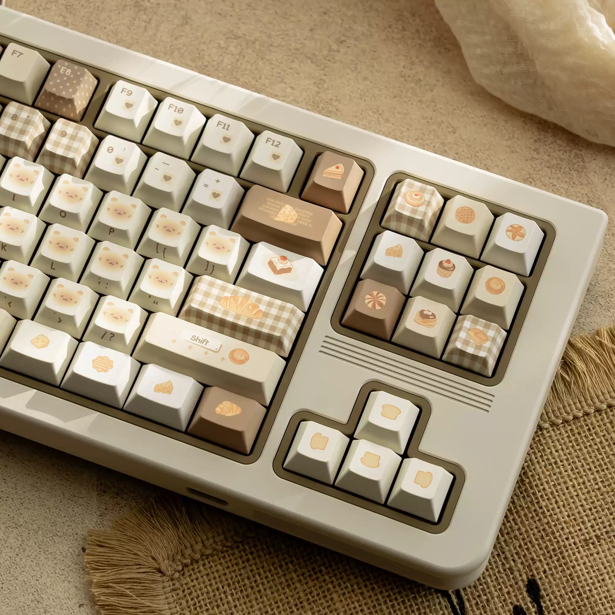A close-up of a mechanical keyboard with cream, beige, and light brown keycaps decorated with cute food illustrations, resting on a textured beige fabric surface.