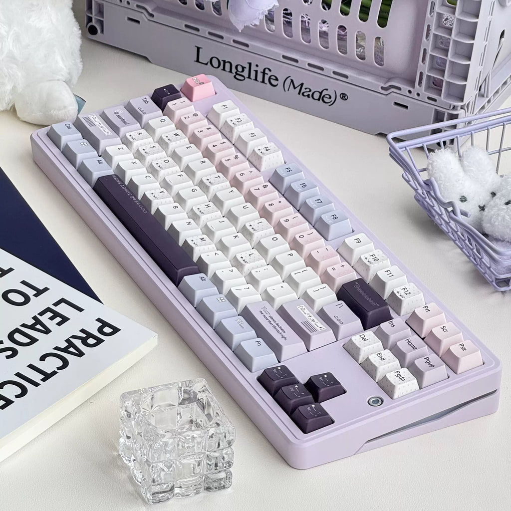 A pastel purple mechanical keyboard with white, pink, and dark gray keycaps, styled on a desk with a plush toy, flowers, and a basket in the background.