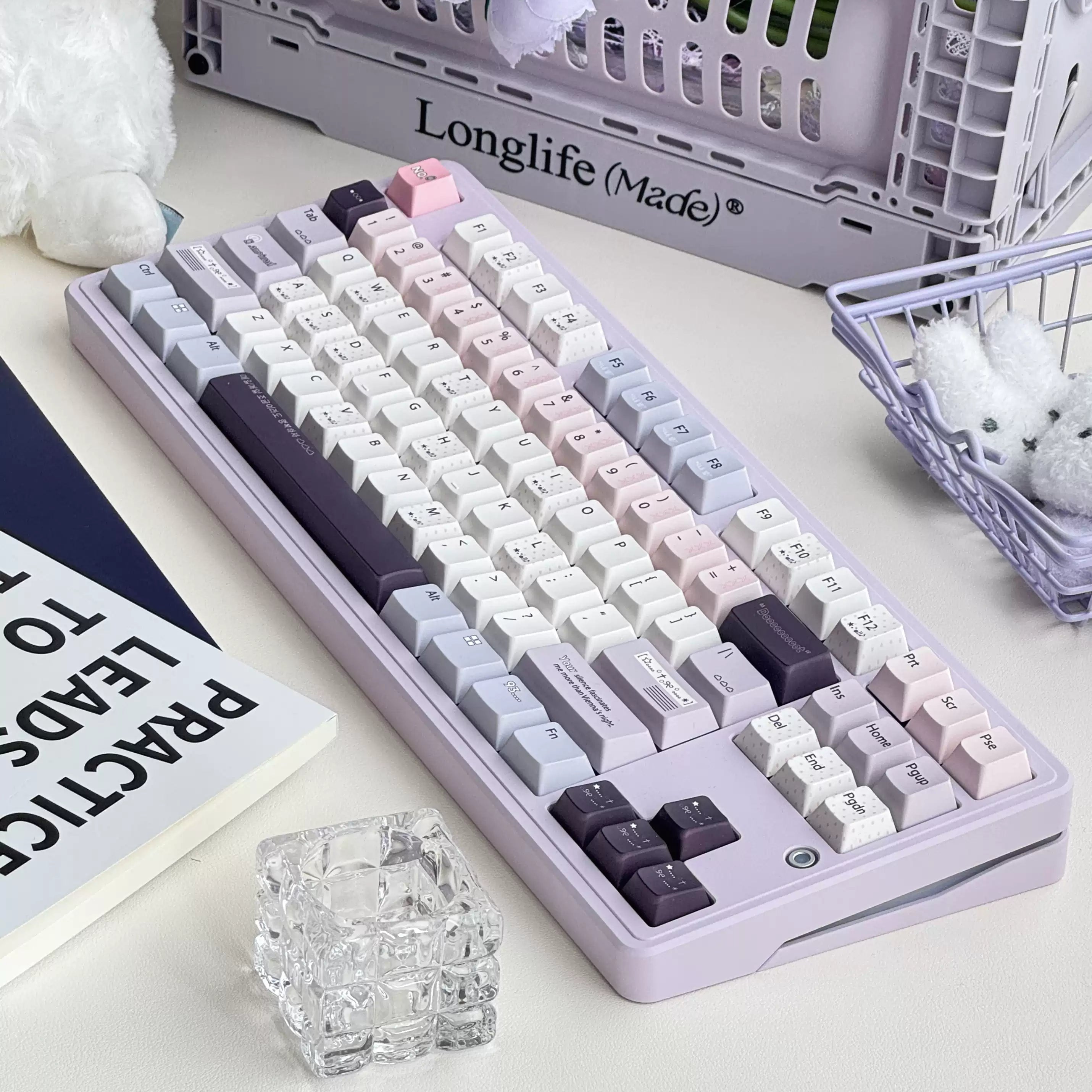 A pastel purple mechanical keyboard with white, pink, and dark gray keycaps, styled on a desk with a plush toy, flowers, and a basket in the background.