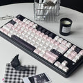 A mechanical keyboard with pastel pink, white, and black keycaps arranged on a round white table, surrounded by desk accessories including a candle, pen, notepad, clips, and a small black-and-white check cloth.