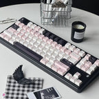 A mechanical keyboard with pastel pink, white, and black keycaps arranged on a round white table, surrounded by desk accessories including a candle, pen, notepad, clips, and a small black-and-white check cloth.