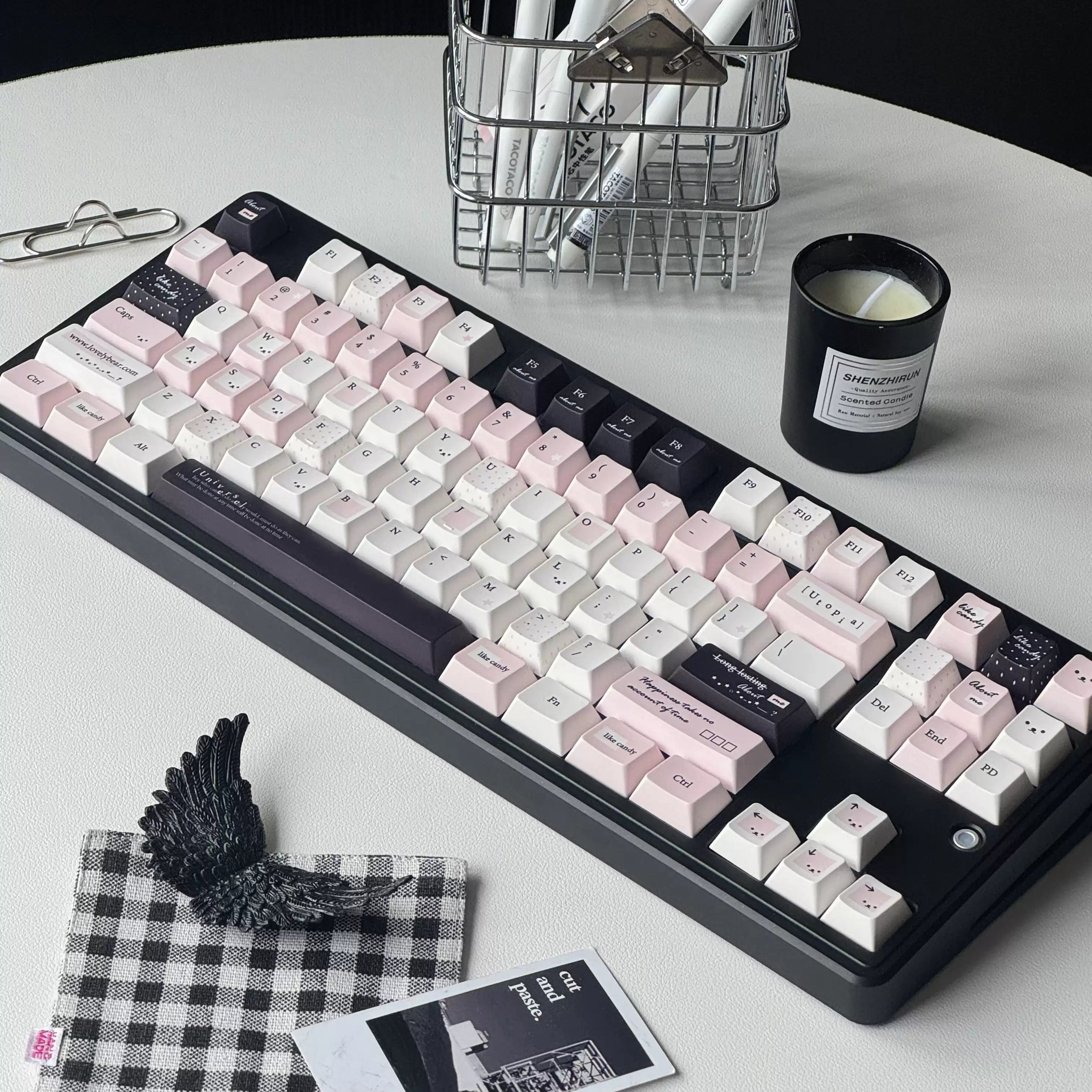 A mechanical keyboard with pastel pink, white, and black keycaps arranged on a round white table, surrounded by desk accessories including a candle, pen, notepad, clips, and a small black-and-white check cloth.