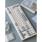 A white mechanical keyboard with minimalist gray and beige keycaps arranged on a light blue-gray desk surface, surrounded by small desk props including a notebook, bottle, and 2026 planner.