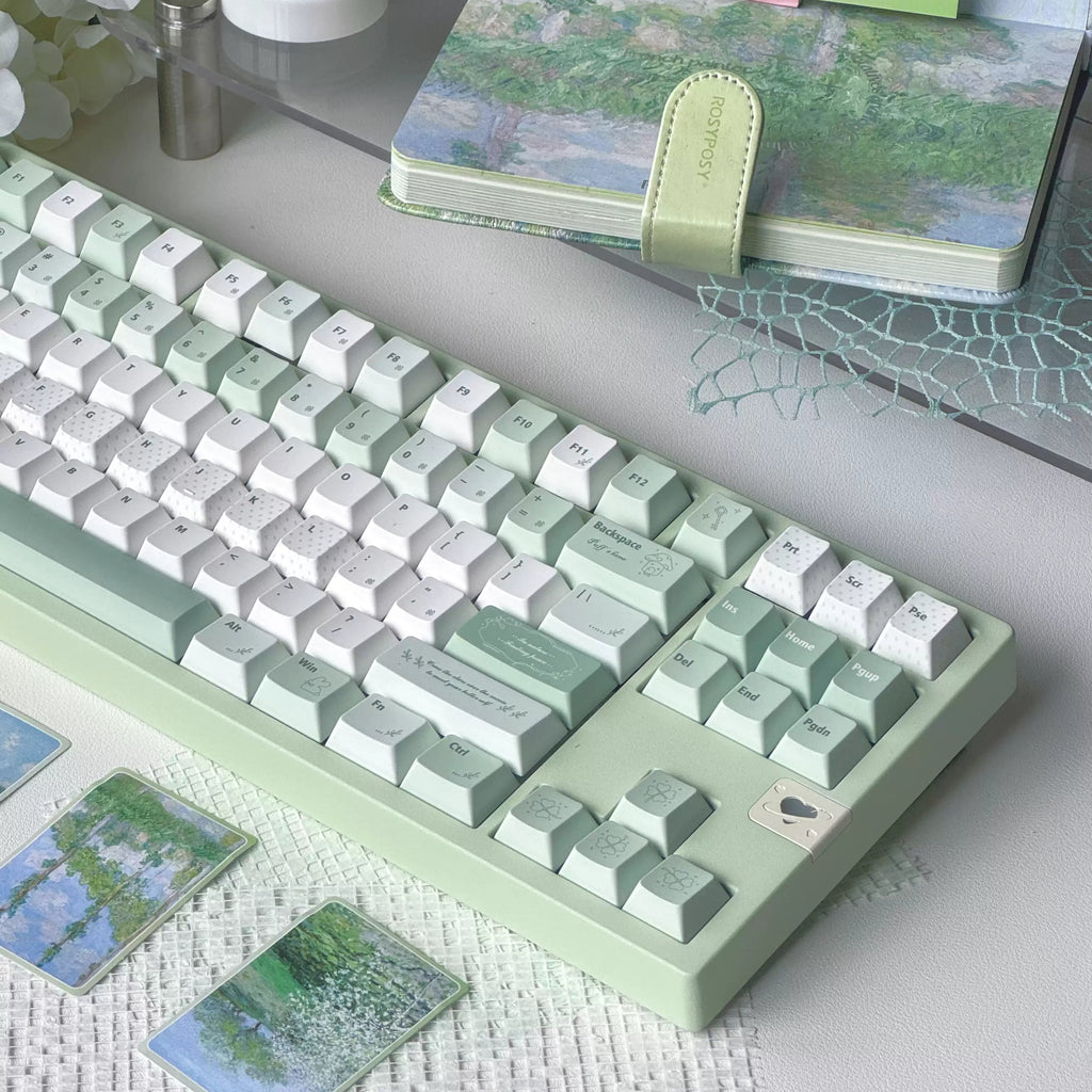 A pastel green and white mechanical keyboard styled on a desk with matching landscape art cards, a notebook, and decorative props.