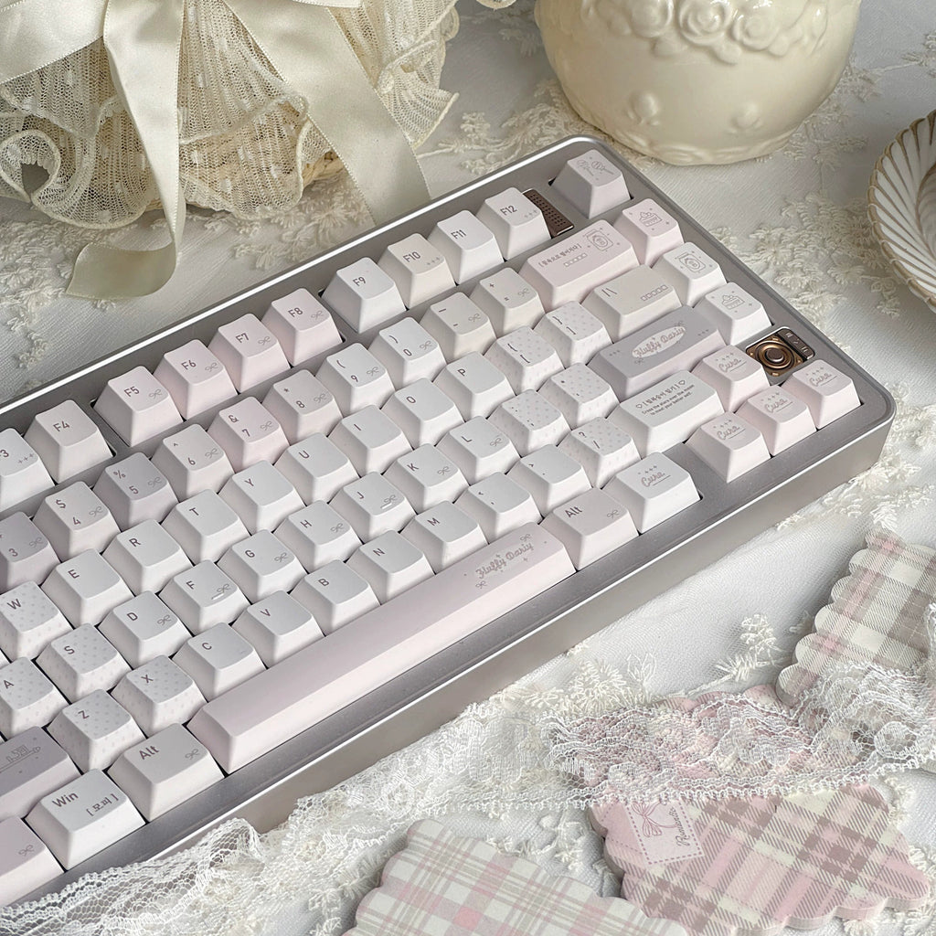 A mechanical keyboard with pale pink and white keycaps and a silver frame, styled on a lace surface with decorative props including a cream vase, a ribboned basket, and pink plaid fabric.