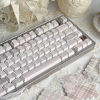 A mechanical keyboard with pale pink and white keycaps and a silver frame, styled on a lace surface with decorative props including a cream vase, a ribboned basket, and pink plaid fabric.