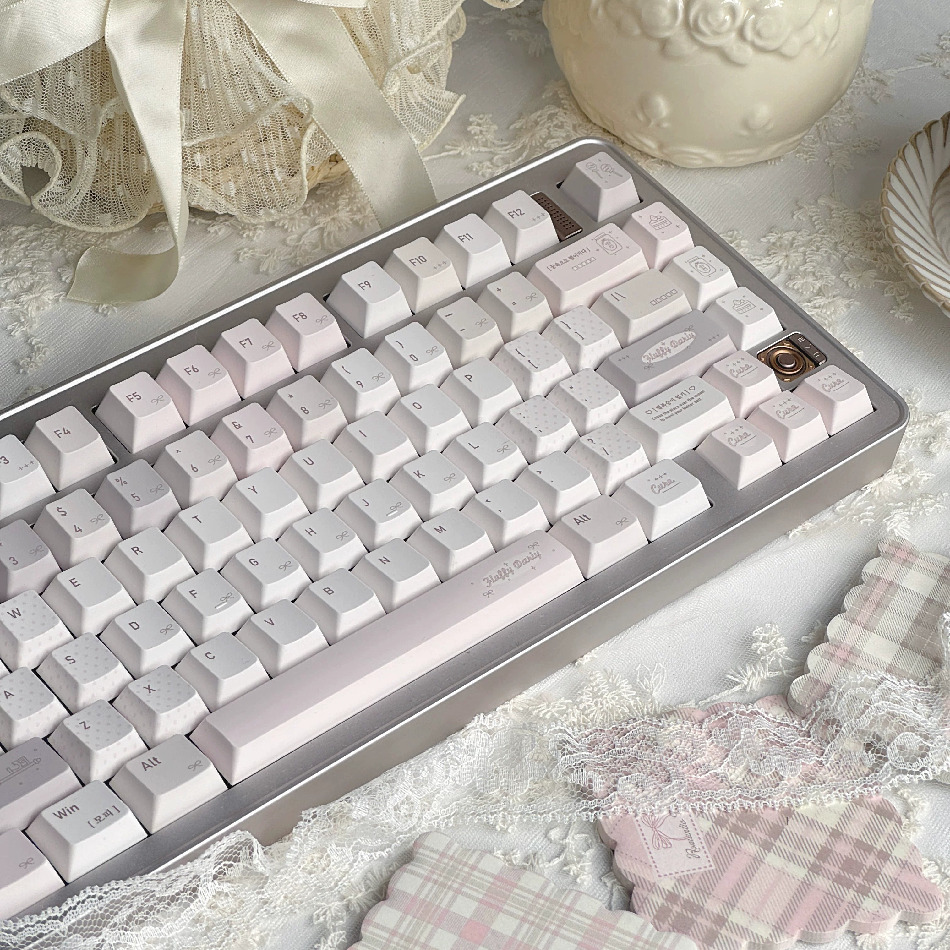 A mechanical keyboard with pale pink and white keycaps and a silver frame, styled on a lace surface with decorative props including a cream vase, a ribboned basket, and pink plaid fabric.