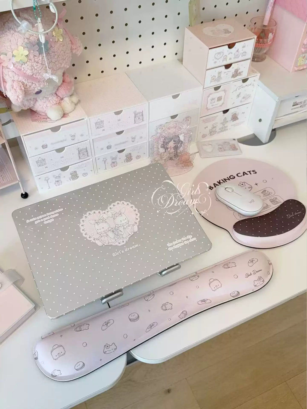 A styled desk setup in pastel pink and white featuring a laptop with a heart-patterned cover, a pink patterned wrist rest, a round mouse pad with a mouse, and a white shelving unit with small drawers in the background.
