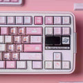 A close-up of a pastel pink mechanical keyboard with Hello Kitty themed keycaps, featuring pink, white, and lavender keys with cute illustrations, set against a matching pink background.