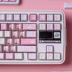 A close-up of a pastel pink mechanical keyboard with Hello Kitty themed keycaps, featuring pink, white, and lavender keys with cute illustrations, set against a matching pink background.