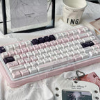 A pastel pink mechanical keyboard with white and dark purple keycaps, styled on a white textured fabric surface with a ceramic mug, cards, and accessories.