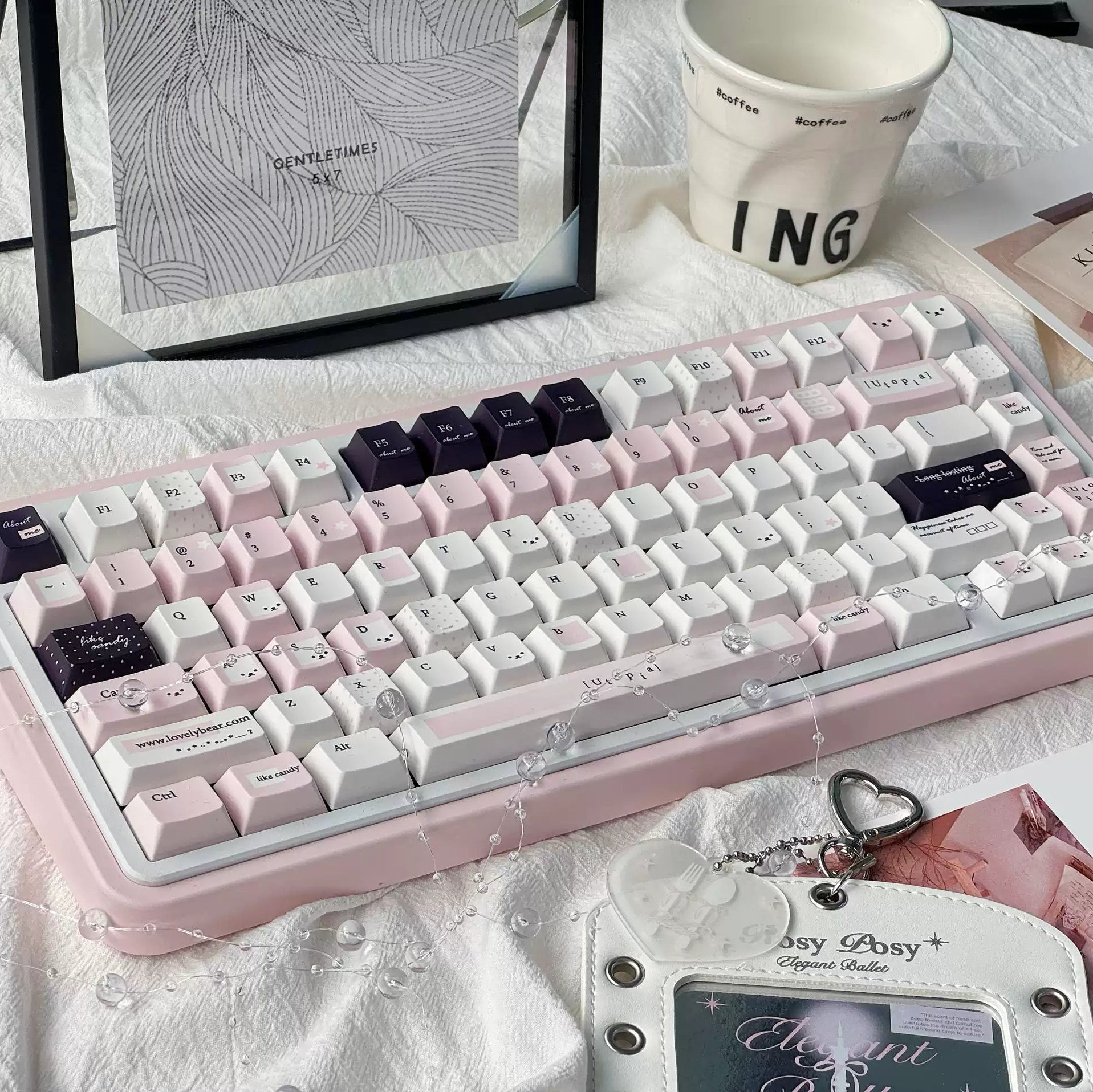 A pastel pink mechanical keyboard with white and dark purple keycaps, styled on a white textured fabric surface with a ceramic mug, cards, and accessories.