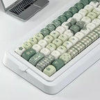 A mechanical keyboard with a white case and pastel green, cream, and white checkered, argyle, and star-patterned keycaps, shown on a white desk with a laptop partially visible in the top left.