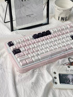 A pastel pink mechanical keyboard with white and light pink keycaps, styled on a white fabric surface with a black picture frame, a white mug, and a small decorative card nearby.